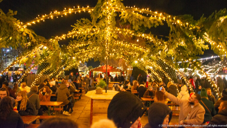 Crowded communal tables beneath arching evergreen branches covered with dense warm string lights.