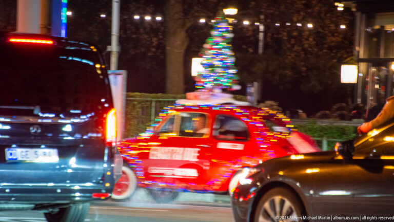 Small red vintage-style car covered in multicolored lights with an illuminated Christmas tree on its roof, moving through traffic.