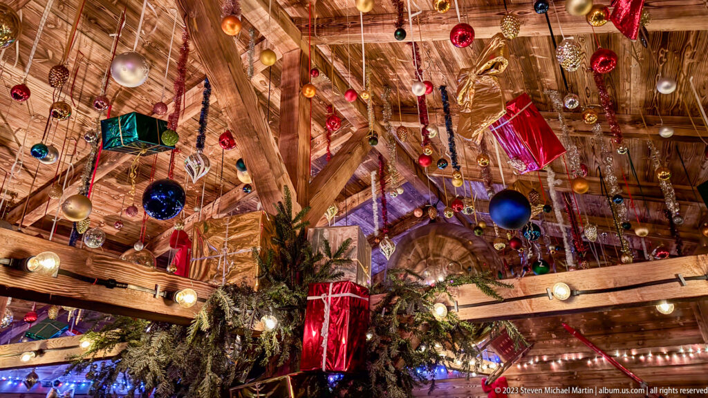 Colorful baubles and suspended gift-box decorations filling multiple timber rafters above evergreen branches and bulbs.