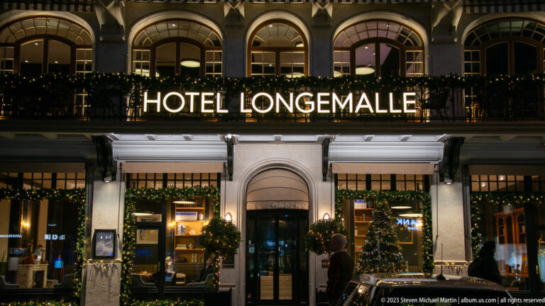 Street-level Hôtel Longemalle entrance with illuminated sign, awnings, greenery, and small Christmas trees.