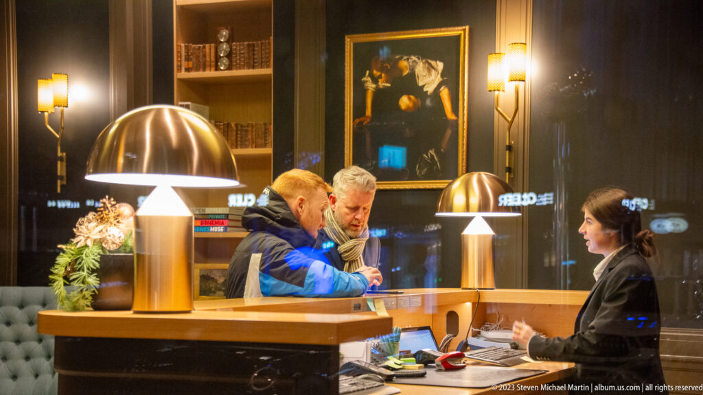 Two men speaking with a receptionist across a wooden hotel desk beneath large brass-toned lamps.