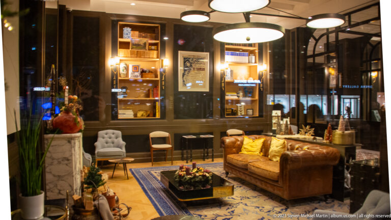 Hotel lounge with brown leather sofa, upholstered chairs, bookshelves, framed art, plants, and circular ceiling lights.