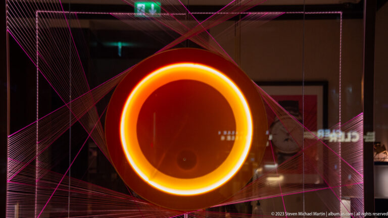 Bright orange circular light ring surrounded by intersecting magenta threads behind reflective glass.