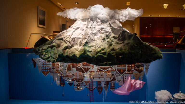 Snow-covered model mountain with clouds above an upside-down miniature town suspended beneath it.