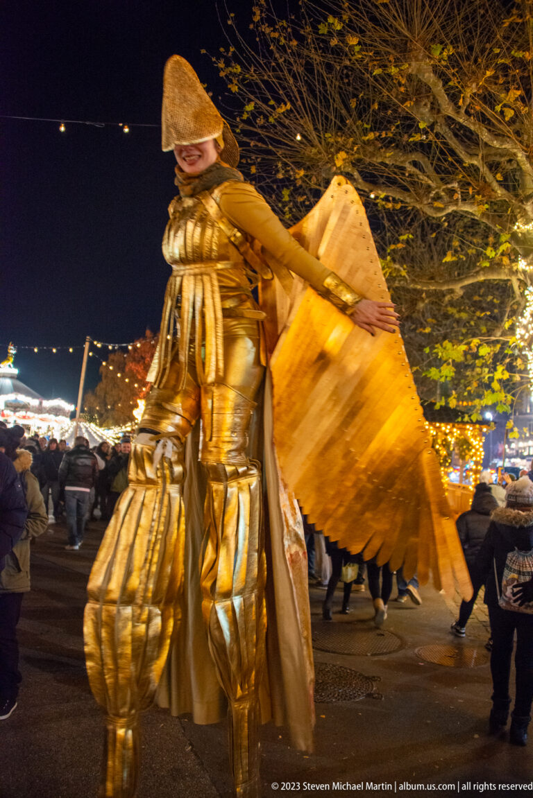 Gold-costumed stilt performer with a large sculptural wing walking through a crowded night market.
