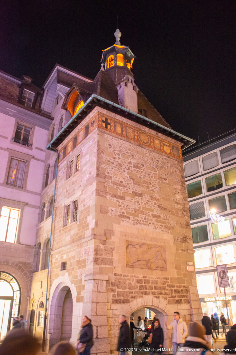 Historic stone gate tower with arched passage, decorated frieze, projecting roof, and pedestrians below at night.