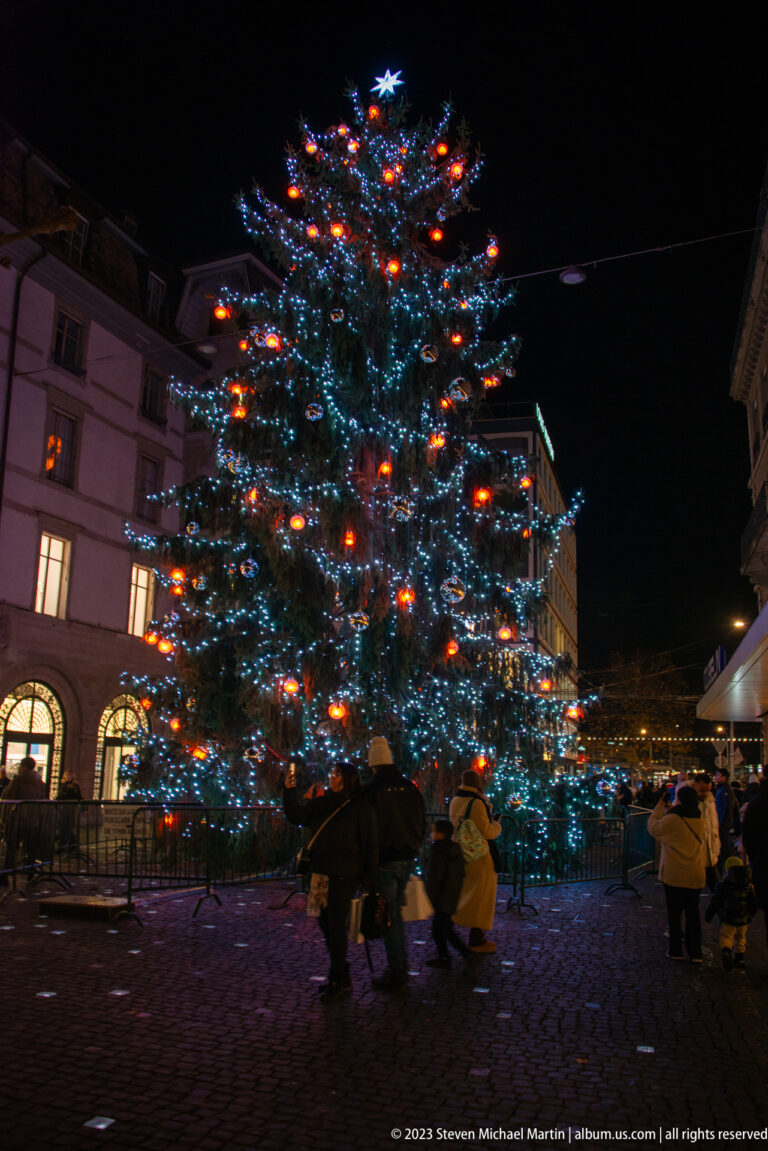 Large outdoor Christmas tree covered in white lights and colorful ornaments, with visitors gathered around its base at night.