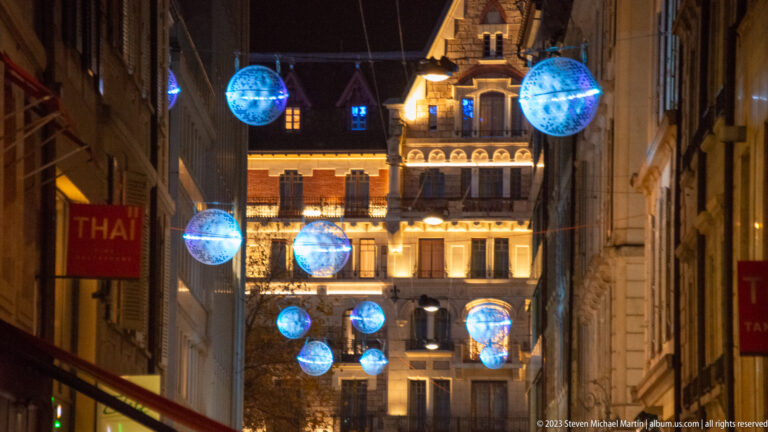 Blue glowing globe decorations suspended above a narrow street leading toward an illuminated historic building.