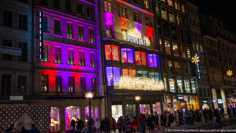 Multi-story city building illuminated in red, purple, and blue, with pedestrians gathered along the storefronts below.