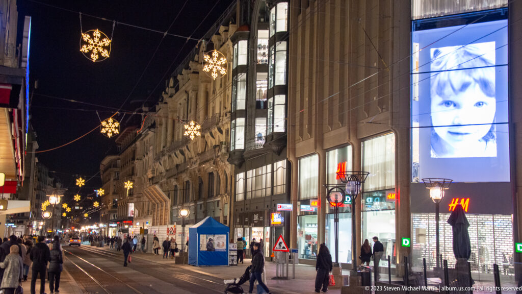 Pedestrians walk beside tram tracks on a shopping street decorated with hanging star-shaped holiday lights at night.