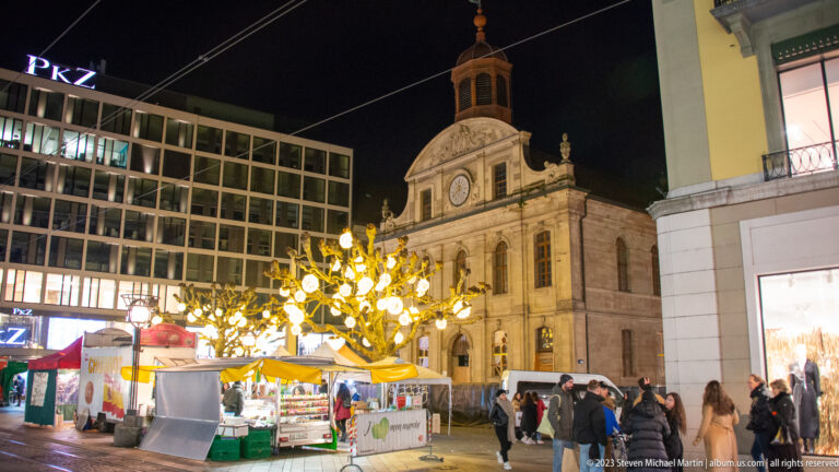Historic pale-stone church with central clock and small bell turret behind market stalls and a glowing decorative tree.