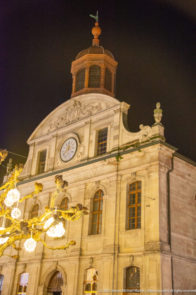 Close nighttime view of a pale stone church façade with central clock, carved ornament, and octagonal roof turret.