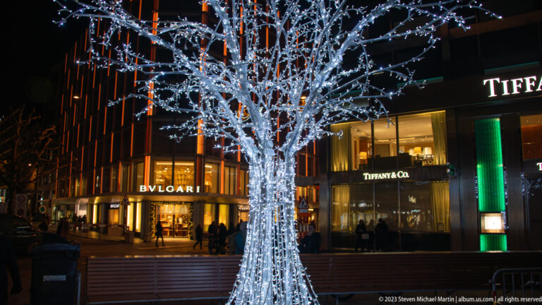 Leafless tree densely wrapped in white lights in front of BVLGARI and Tiffany & Co. storefronts at night.