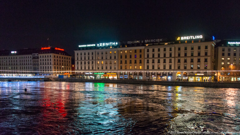 Illuminated Geneva buildings and watch-brand signs reflected in the dark Rhône at night.