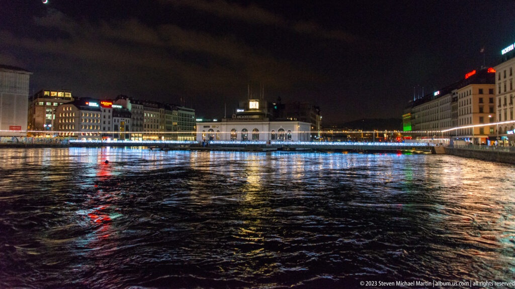 Wide nighttime view along the Rhône with illuminated buildings, a distant bridge, and colored reflections on the water.