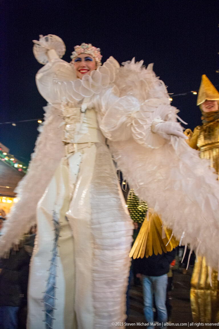 Tall white-costumed stilt performer with feathered forms moving through the night market.