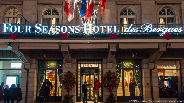 Illuminated Four Seasons Hotel des Bergues entrance with flags, holiday decorations, and pedestrians outside.