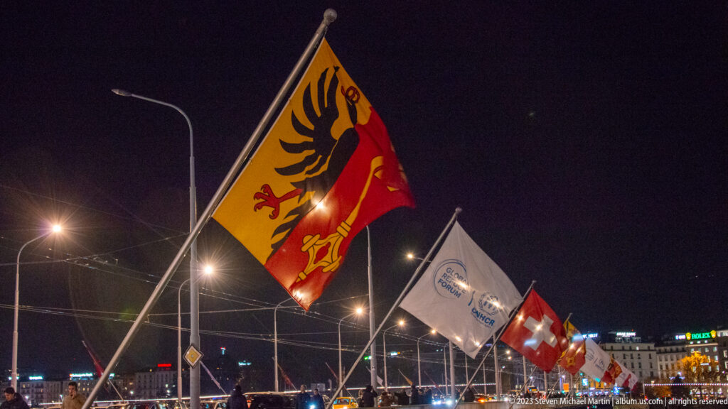Canton of Geneva flag with black eagle and gold key flying beside Swiss and other flags at night.