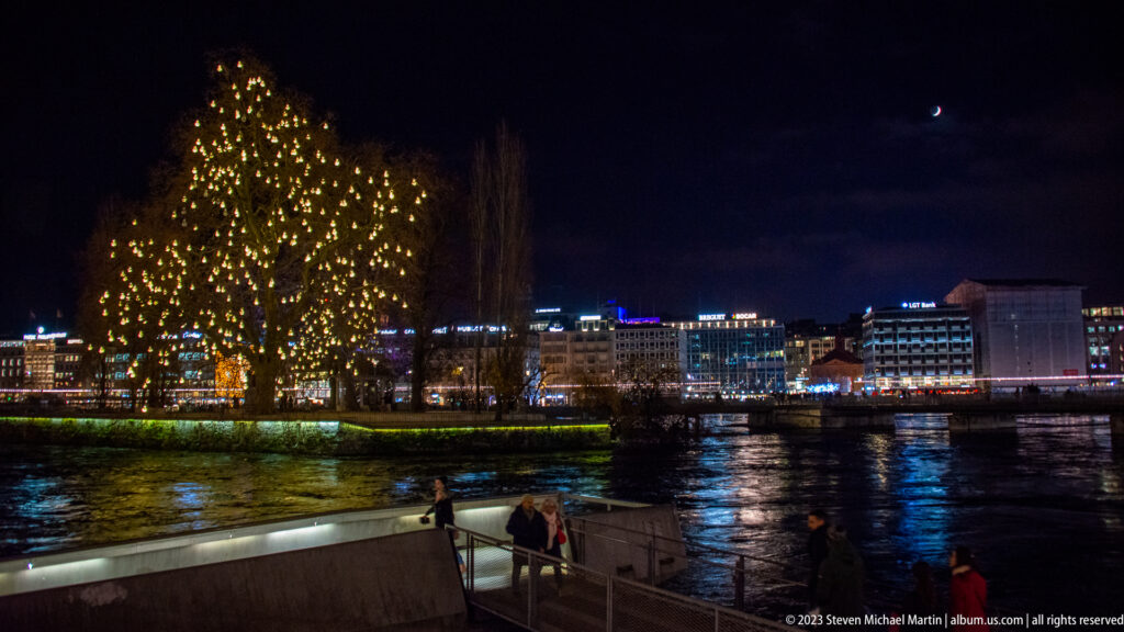 Large riverside tree covered in golden lights beside the Rhône, with city reflections and a crescent moon at night.