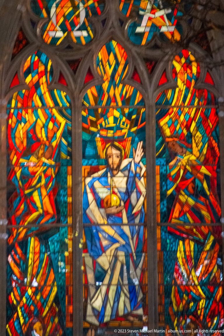 Colorful modern stained glass with a haloed Christ figure holding a chalice, framed by dark Gothic-style tracery.