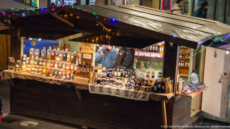 Wooden Christmas-market stall lined with bottles of spirits and liqueurs, price cards, and a vendor behind the counter.