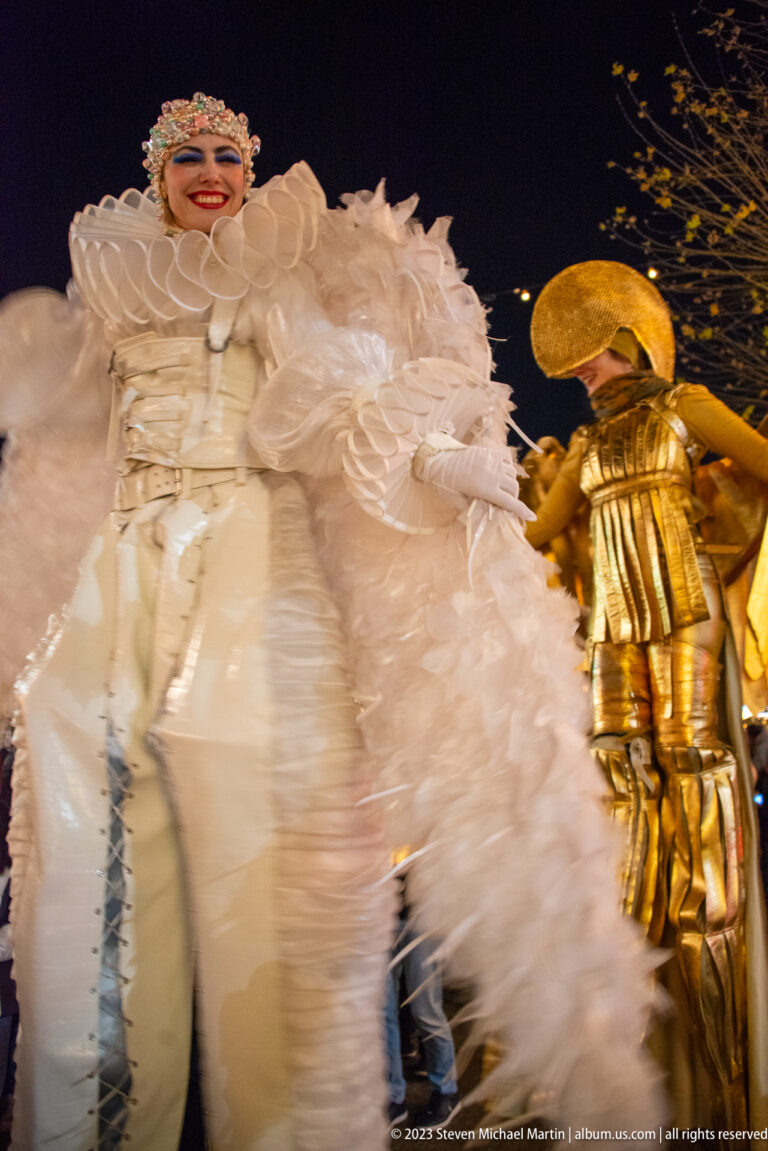 Close low-angle view of a white feathered stilt performer with a gold-costumed performer behind.