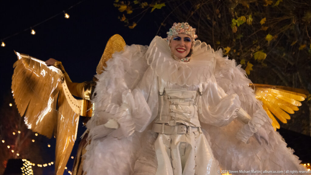 Smiling performer in an elaborate white feathered costume and jeweled headpiece, with golden wing-like forms behind.