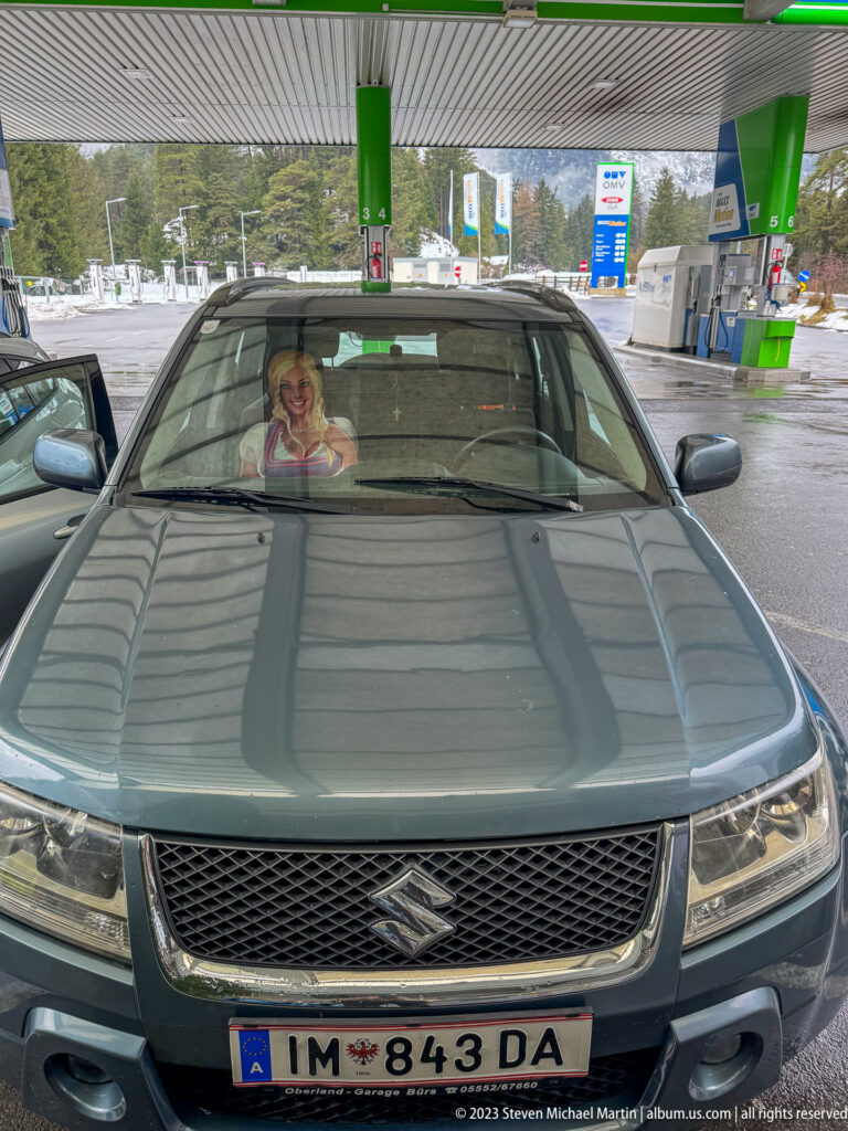 Front of a dark Suzuki beneath a service-station canopy, with snowy mountains beyond and a printed female figure behind the windshield.