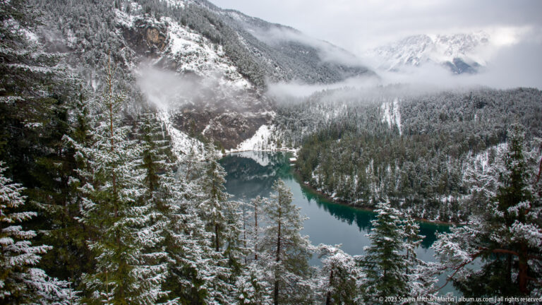 Elevated view of an elongated turquoise lake surrounded by snowy conifer forest and cloud-covered mountains.