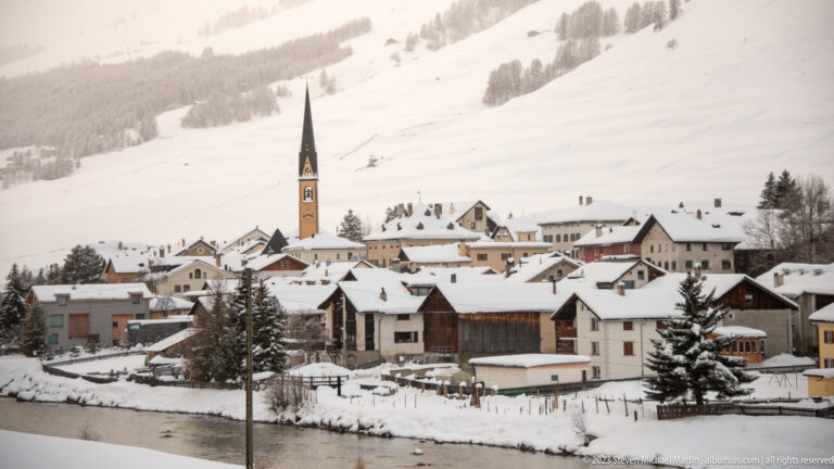 Snow-covered alpine village clustered around a tall dark church spire beneath broad white mountain slopes.