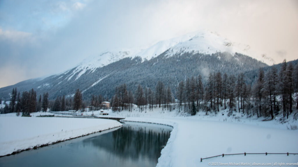 Calm blue-green river bending between deep snowbanks below a forested, cloud-wrapped mountain.