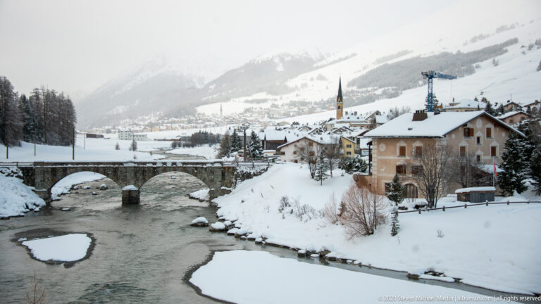 Two-arched stone bridge crossing a snowy river beside village houses and a distant church spire.