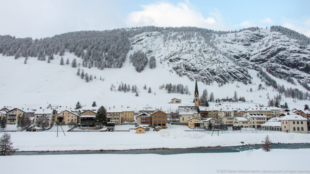 Snow-covered alpine village with colorful houses and a slender church tower beneath a steep rocky mountainside.