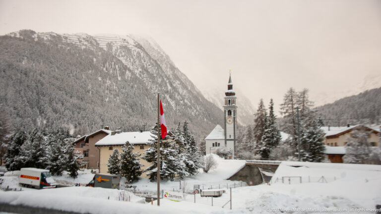 White alpine church with tall bell tower, Swiss flag, snowy buildings, and forested mountains behind.