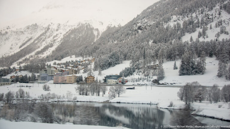 Snowy alpine settlement reflected in calm water beneath steep forested mountain slopes.