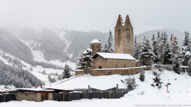 Stone church on a snowy rise beside a tall ruined tower with two pointed wall remnants.
