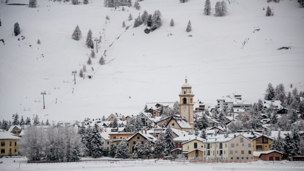 Snow-covered alpine village with a square church tower and ski slopes rising behind clustered houses.