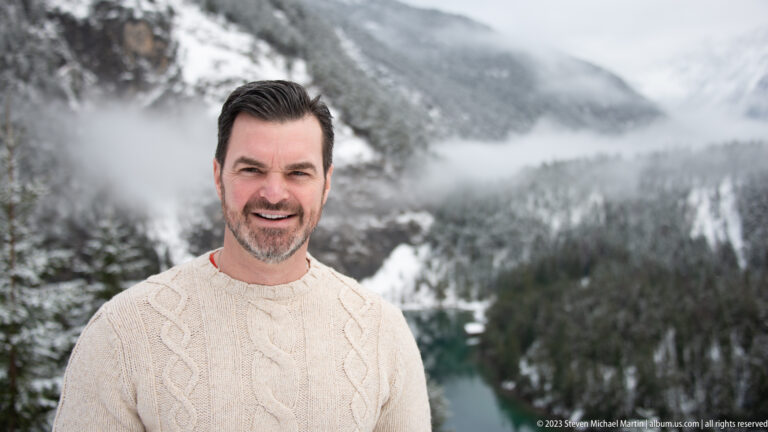 Smiling dark-haired man in a cream cable-knit sweater with a snowy Alpine lake and forest blurred behind him.