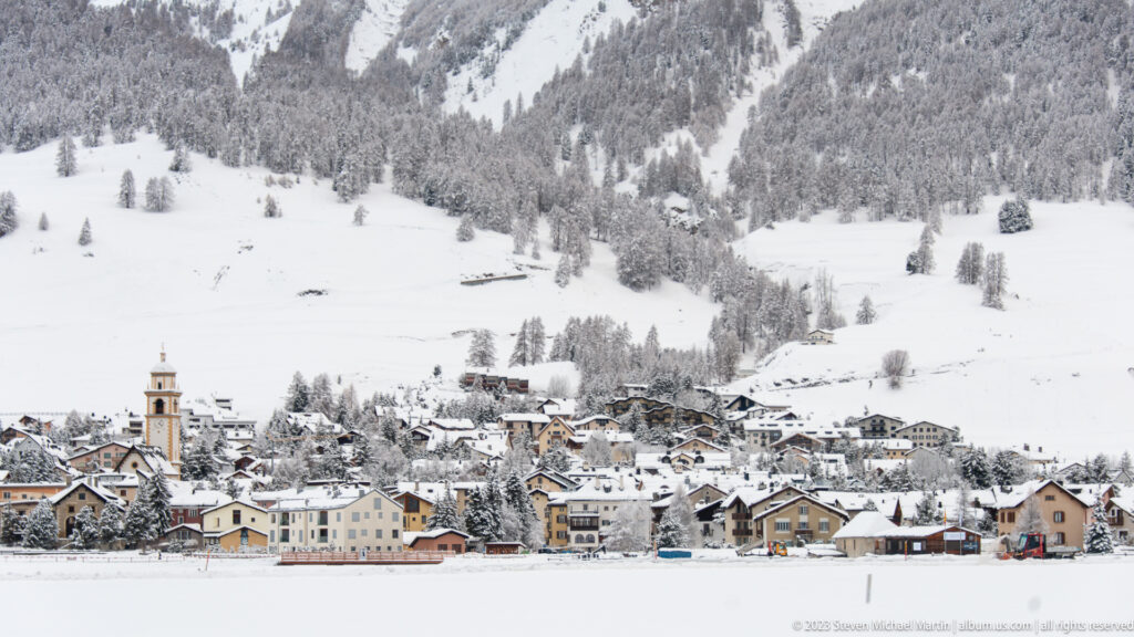 Wide snow-covered alpine village beneath forested mountain slopes and open white fields.