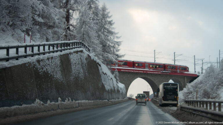Red electric passenger train crossing a stone arch above cars on a wet snow-lined mountain road.
