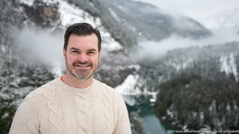 Close portrait of a smiling dark-haired man in a cream sweater, with snowy forest and turquoise lake blurred behind.