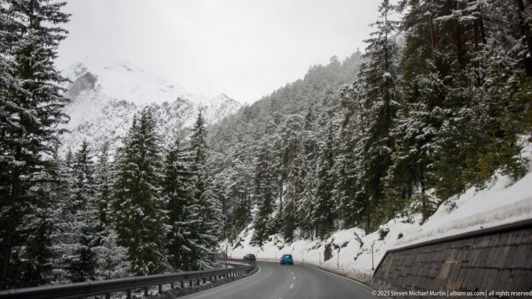 Two-lane mountain highway with cars, guardrails, snowy conifers, and steep cloud-covered Alpine slopes.