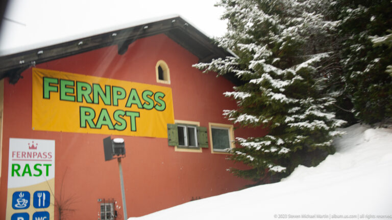 Orange-red roadside building in snow with large yellow-and-green 'FERNPASS RAST' lettering and service signs.