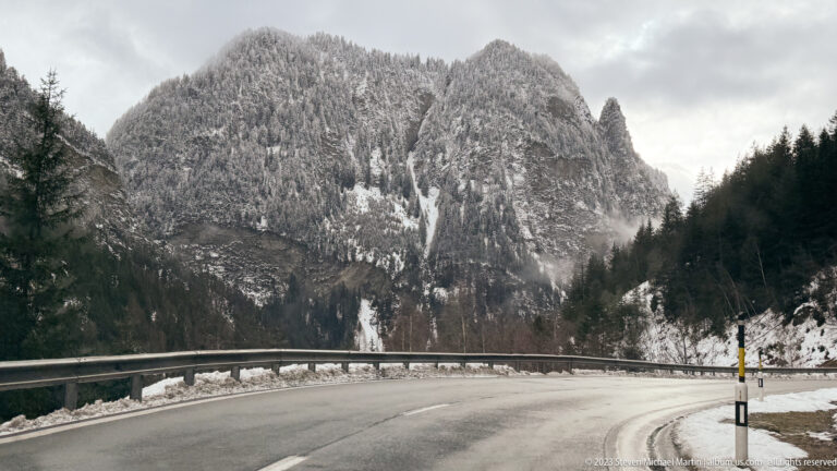 Curving wet mountain road with guardrail below steep snow-dusted cliffs and forested slopes.