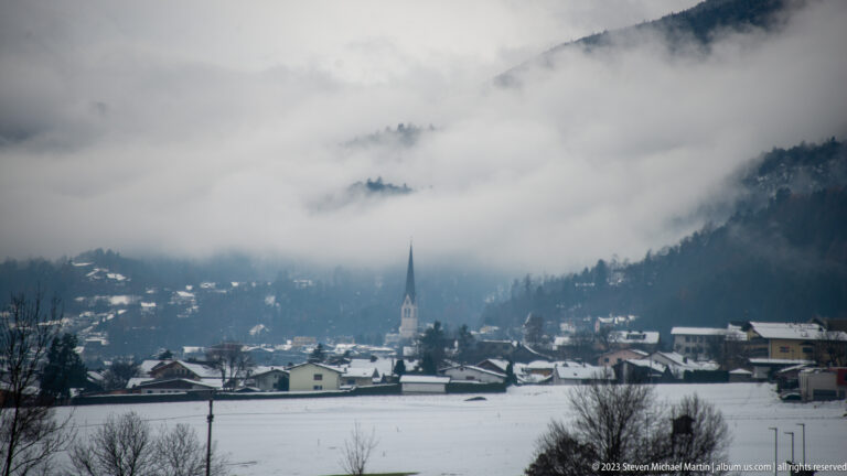 Snowy Alpine village with a tall church spire beneath a mountain largely hidden by low cloud.