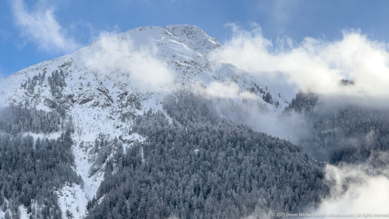 Broad snowy mountain above dense frosted conifer forest, with a cloud bank crossing the upper slope.