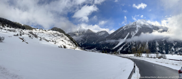 Narrow dark road crossing a broad snowy valley toward rugged mountains under broken blue sky and cloud.