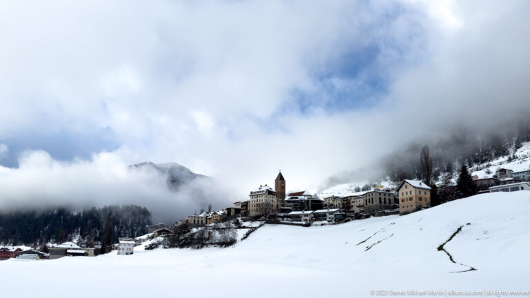 Small snowy Alpine village on a rise with a prominent square tower and cloud-covered mountains behind.