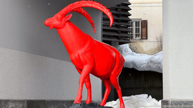 Bright red sculpture of an Alpine ibex with long curved horns standing beside a building in snow.