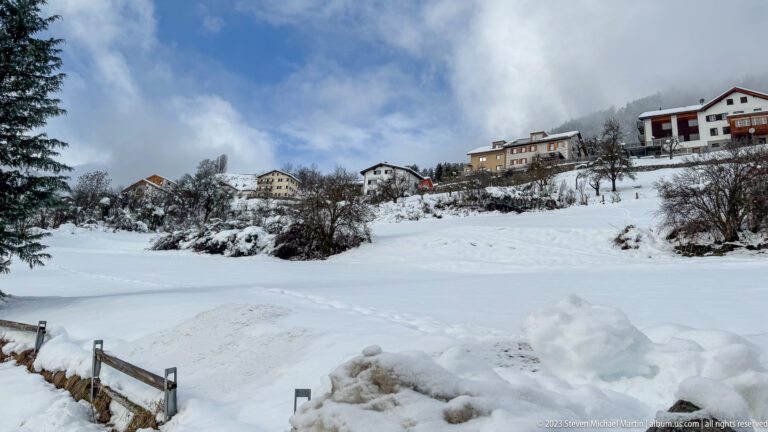 Scattered houses on a snowy slope above a broad white field, with bare shrubs and blue sky.